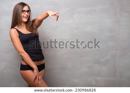 Half-length portrait of sexy smiling dark -haired young woman in nice eyeglasses wearing black vest and shorts standing aside showing her perfect figure pointing at someone