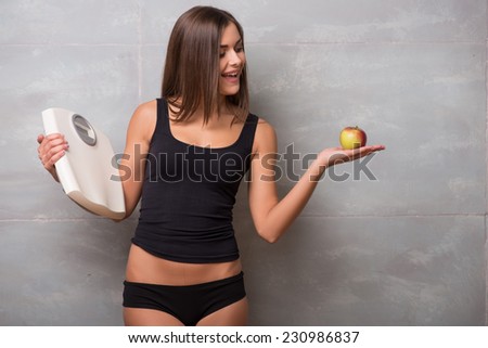 Half-length portrait of lovely smiling dark -haired young woman wearing black vest standing aside holding in one hand very delicious ripe apple and in another electronic balance