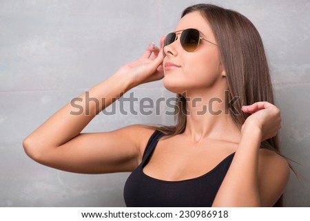 Half-length portrait of beautiful smiling sexy dark haired young woman wearing great sunglasses and black vest standing aside looking out for someone