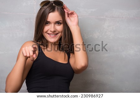 Half-length portrait of beautiful smiling sexy dark -haired young woman wearing black vest keeping her wonderful sunglasses on the head having fun pointing at something