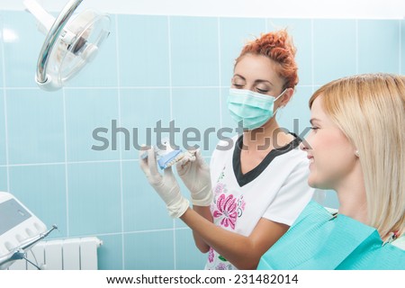 Half-length portrait of young fair-haired lovely smiling girl sitting on the dentist chair looking at the doctor wearing a mask which is showing her false tooth