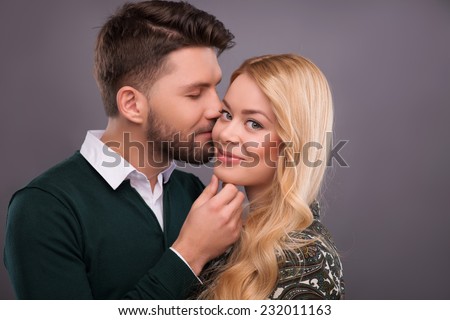 Half-length portrait of handsome young man wearing white shirt and dark-green cardigan standing hugging his beautiful fair-haired girlfriend wanted to kiss her