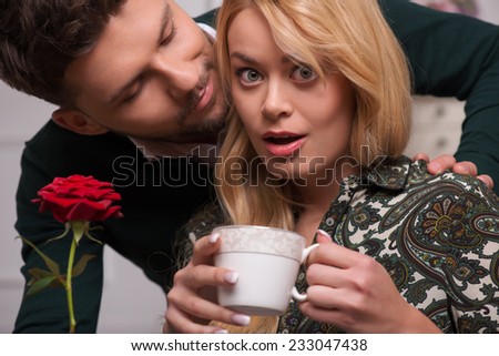 Selective focus on the beautiful surprised fair-haired young woman wearing blouse and jeans looking happy at her boyfriend giving her wonderful red rose