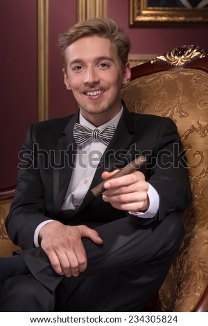 Half-length portrait of handsome young fair-haired man wearing classic costume sitting relaxing on the comfortable chair with his favorite cigar satisfied with his life