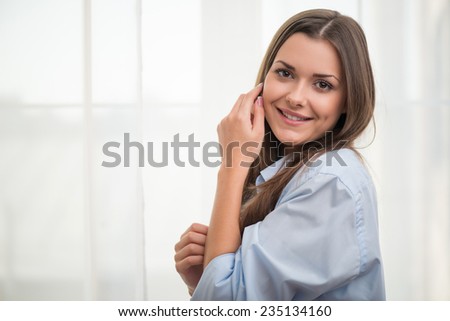 Half-length portrait of dark-haired beautiful smiling girl wearing blue shirt standing aside touching her neck looking at us thinking about wonderful last date