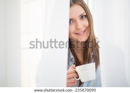 Selective focus on the dark-haired beautiful smiling girl waking up in the morning standing near the window looking out of the white curtains holding a cup of hot delicious coffee