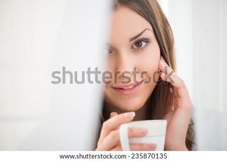 Selective focus on the dark-haired beautiful smiling girl waking up in the morning standing near the window looking out of the white curtains touthing her face holding a cup of hot delicious coffee