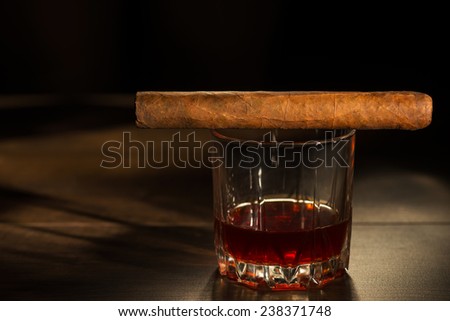 Selective focus on the Cuban cigar lying on the glass of whiskey that standing on the wooden table