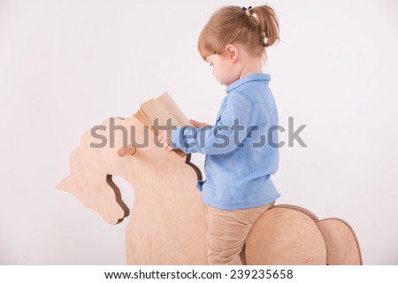 Half-length portrait of little lovely girl wearing blue shirt and brown pants riding on the wooden horse reading a book. Isolated on the white background