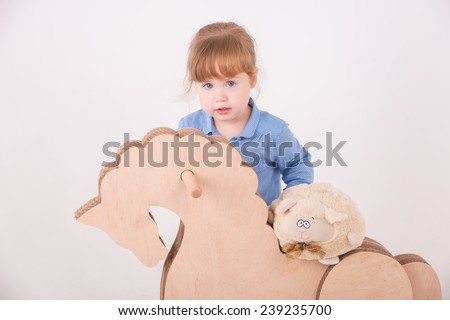 Half-length portrait of little lovely girl wearing blue shirt and brown pants leaning on the wooden horse holding a soft toy. Isolated on the white background