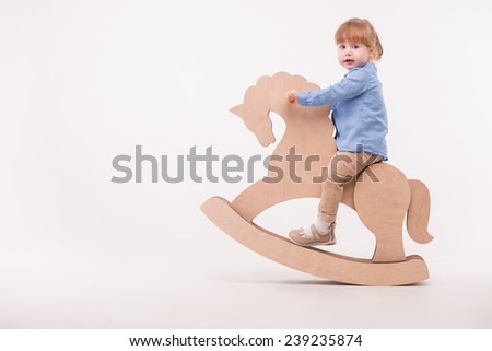 Full-length portrait of little lovely smiling girl wearing blue shirt and brown pants swaying on the wooden toy horse. Isolated on the white background