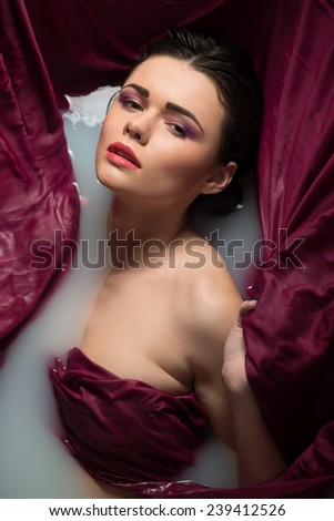 Half-length portrait of beautiful sexy dark-haired woman lying in the bath covering herself with the wine-colored cloth looking at us seductively. Top view