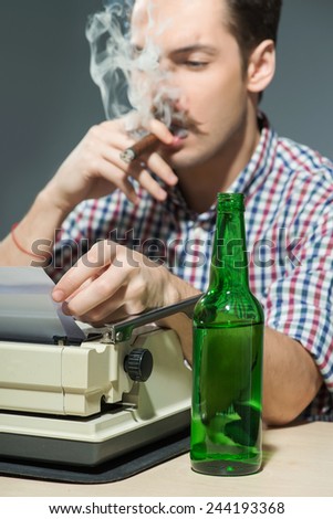 Author at work. Close-up of creative young author working at the typewriter, smoking cigar and drinking alcohol while sitting at his working place against grey background