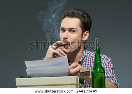 Author at work. Creative young author working at the typewriter, smoking cigar and drinking alcohol while sitting at his working place against grey background