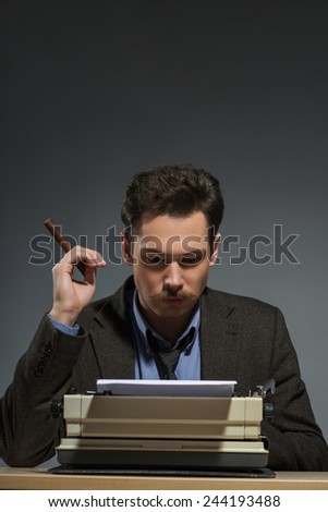 Author at work. Close-up of confident young author working at the typewriter and smoking cigar while sitting at his working place against grey background