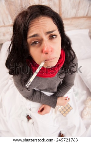 Sick woman with thermometer. Top view image of young frustrated woman in knitted scarf holding thermometer in her mouth while sitting against wooden wall surrounded by medicines