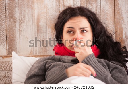 Sick woman caught a cold. Closeup image of frustrated young woman with red nose lying in bed with thick scarf and sneezing into tissue