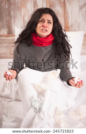 Sick and hopeless. Young woman with flu wearing thick scarf raising her hands with tissues in despair while sitting in bed on country house background