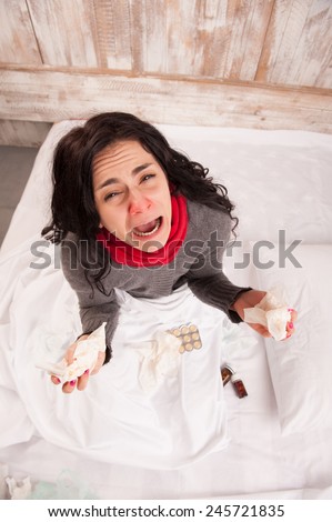 Desperate and sick. Top view image of young sick woman crying while sitting on bed with pills and tissues against wooden wall