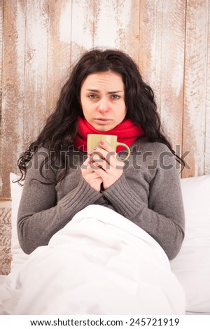 Sick woman with cup of tea. Closeup image of young frustrated woman in knitted scarf holding a cup of tea while sitting in bed against wooden wall