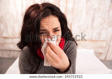 Caught a cold. Closeup image of young sick woman blowing her nose while sitting on bed against wooden wall