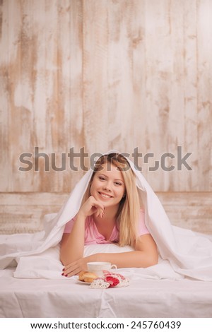 Happy valentines day. Young woman covered with blanket having breakfast with coffee and croissants served to bed and holding rustic gift hearts
