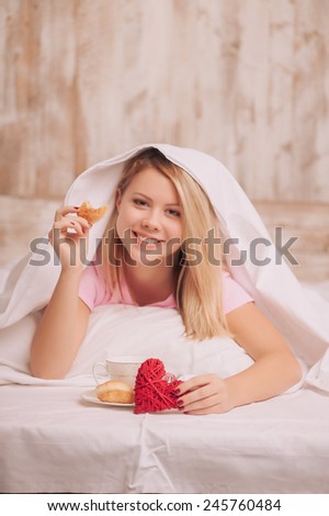 Beauty having breakfast in bed. Young woman sitting in bed with coffee and eating croissants served to bed due to valentines day, selective focus on rustic gift hearts
