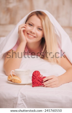 Breakfast in bed. Closeup image of young woman covered with blanket having french breakfast with coffee and croissants served to bed due to valentines day, selective focus on rustic gift hearts