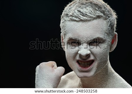 Frozen and mystical. Conceptual photo of young aggressive evil looking man with a face covered with ice powder showing fist and evil grin while standing isolated on black background