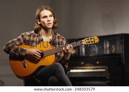 Creative soul. Handsome young sensual man in checked shirt playing acoustic guitar while sitting against the loft room with a vintage piano