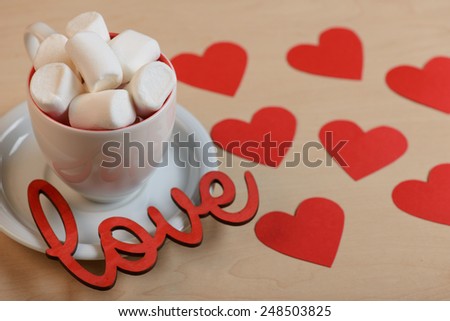 Love is in the air. Top view of the cup full of white marshmallows standing on the table with decorative red hearts and love message