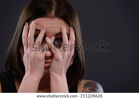 Scared victim. Closeup portrait of terrified teenage girl looking through fingers while standing on dark background