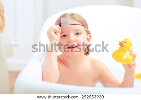 Happy days of childhood. Top view image of a cute little girl in sleep mask taking a bath and playing with rubber ducks while sitting in a luxurious bathtub