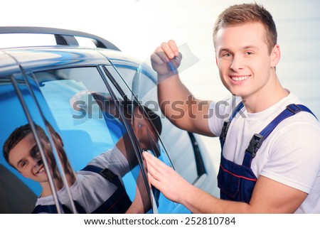 Welcome to our car service station. Closeup image of a handsome car mechanic attaching tinting foil to car window and smiling at camera in specialized service station