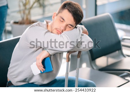 Exhausted traveler. Tired handsome man in casual clothing sleeping on his luggage while sitting on the rows of chairs in the airport lobby with a ticket and passport in hand