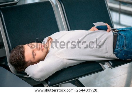 Time to nap. Tired handsome man in casual clothing sleeping on the rows of chairs in the airport lobby with a ticket and passport in hand