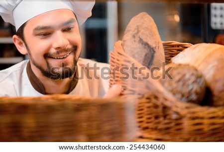 Proud of his baked goods. Cropped image of a handsome young man in uniform looking at a basket with baked goods in the shop window in bakery shop