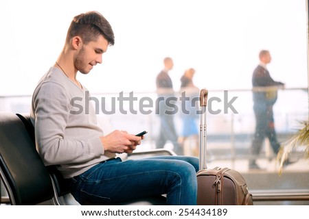 Texting he is fine. Portrait of handsome smiling man in casual wear holding his luggage and messaging through his mobile phone while sitting in the hall of the airport