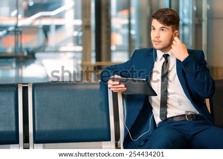 Always on the go. Portrait of handsome smiling businessman in formalwear holding a digital tablet with earphones while sitting in the airport in business lounge