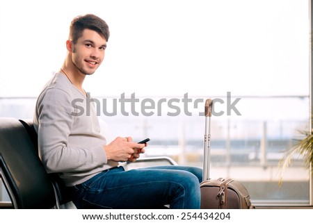 Texting he is fine. Portrait of handsome smiling man in casual wear holding his luggage and messaging through his mobile phone while sitting in the hall of the airport