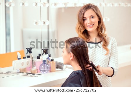 I love my job. Beautiful hairdresser doing hairstyle to her client and smiling at camera while standing on the background of a professional hairdressing salon
