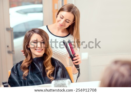 Going for big curls. Mirror reflection of a young beautiful woman discussing hairstyling with her hairdresser while sitting in the hair salon and getting her hair done with hair iron