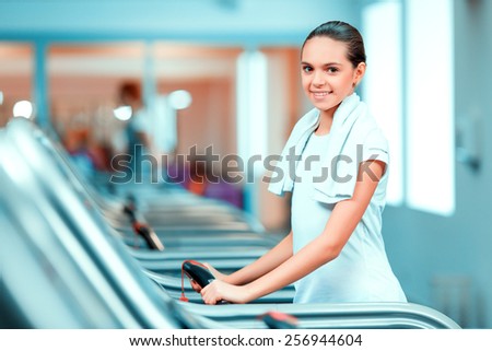 Cardio training. Side view of beautiful teen girl n in sports clothing and towel on her shoulders smiling after her workout on a treadmill in the gym