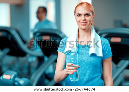 Sporty mom. Attractive mature woman in sports clothing holding bottle with water while standing in health club with a towel on her shoulders