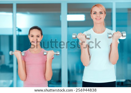 Exercising with dumbbells. Beautiful mature woman and her teenage daughter in sports clothing exercising with dumbbells and smiling while standing against glass wall in the gym