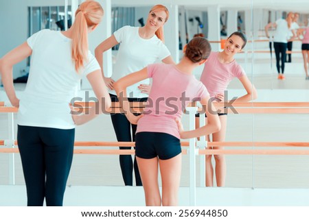 Stretching is a great start of the day. Beautiful mature woman and her teenage daughter in sports clothing warming up before the workout while standing against mirror in health club