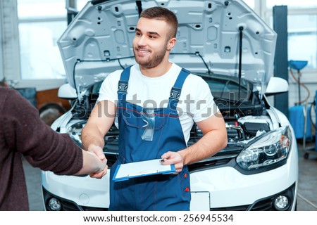 Confident mechanic. Portrait of a handsome car mechanic shaking hands with his client and holding his clipboard in the car repair station
