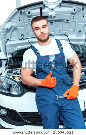 Following my passion to cars. Portrait of a smiling handsome mechanic in uniform posing by the car at car service station holding repair tools