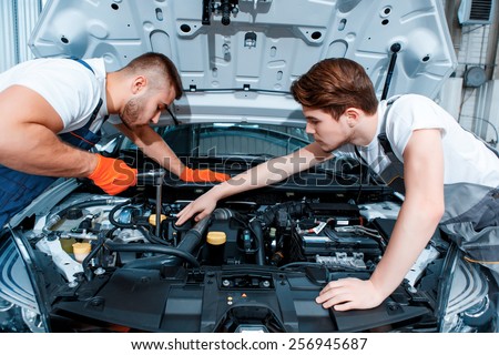 They can fix anything. Two handsome car mechanics in uniform checking the engine under hood in the car service station