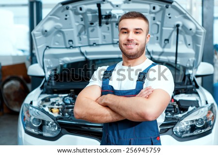 Welcome to our service station. Portrait of a smiling handsome mechanic in uniform posing by the car at car service station with his hands crossed